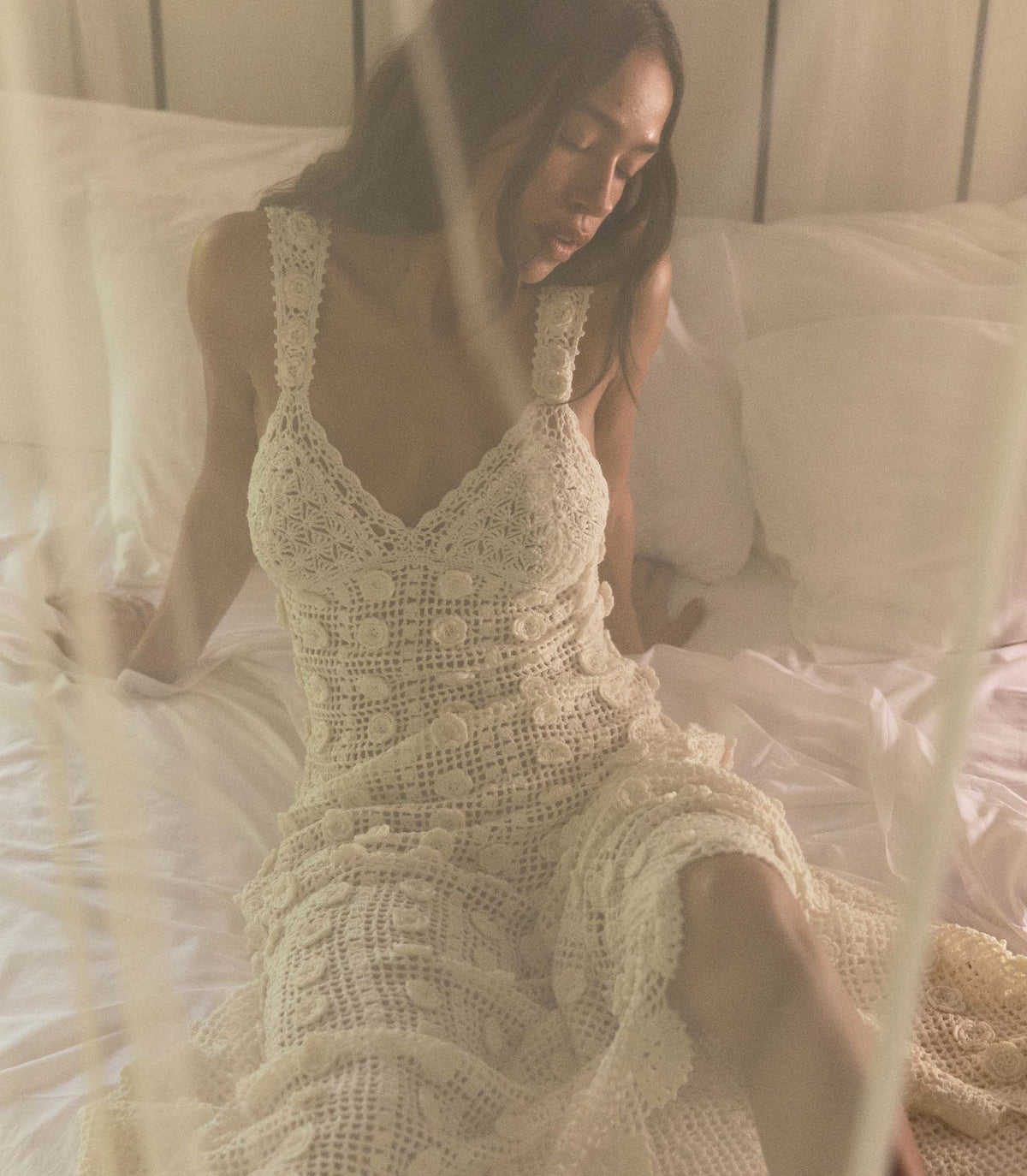 Woman wearing a white crochet dress sitting on a bed.