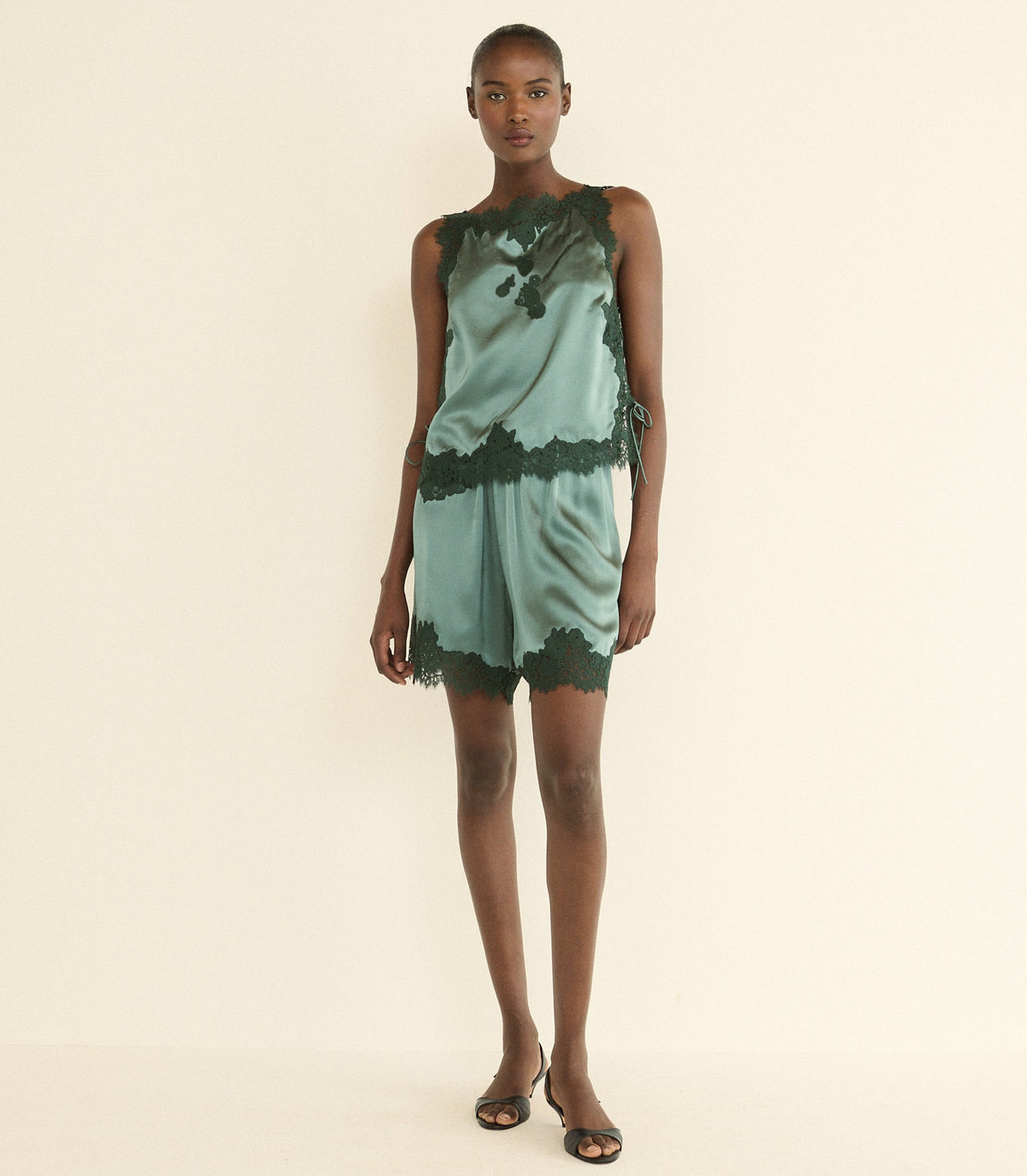 A model stands against a plain cream studio background wearing a sleeveless green satin top with dark green lace trim and a dark green abstract print.