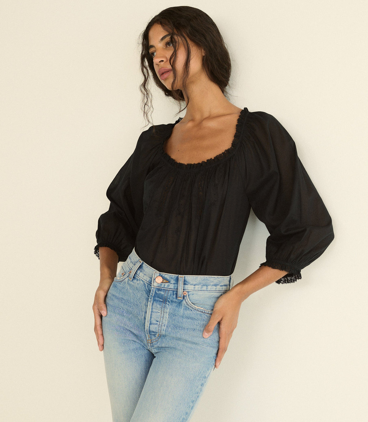 A woman wearing a black puff-sleeve blouse with a square neckline and scalloped trim, paired with light blue jeans, posing against a plain off-white background.