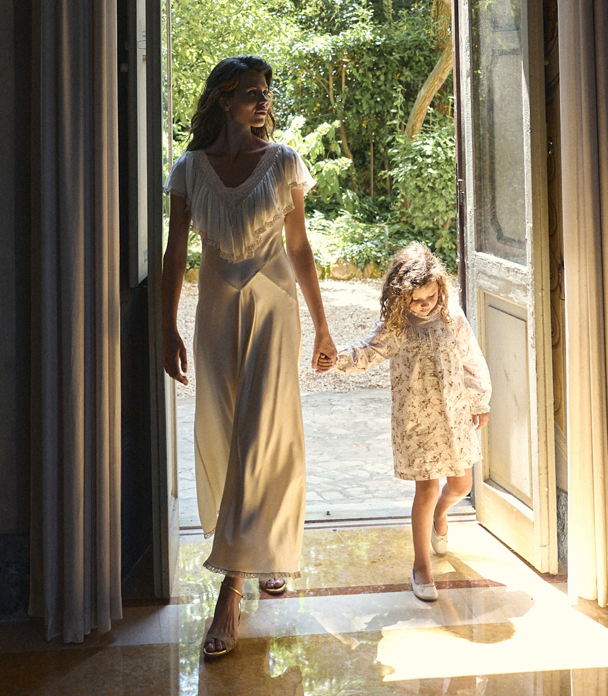 A woman and a young girl walk hand in hand out of a sunlit room through open doors, wearing matching light-colored dresses. The woman wears a cream satin slip dress with fringe at the neckline, and the girl wears a matching cream floral print dress.