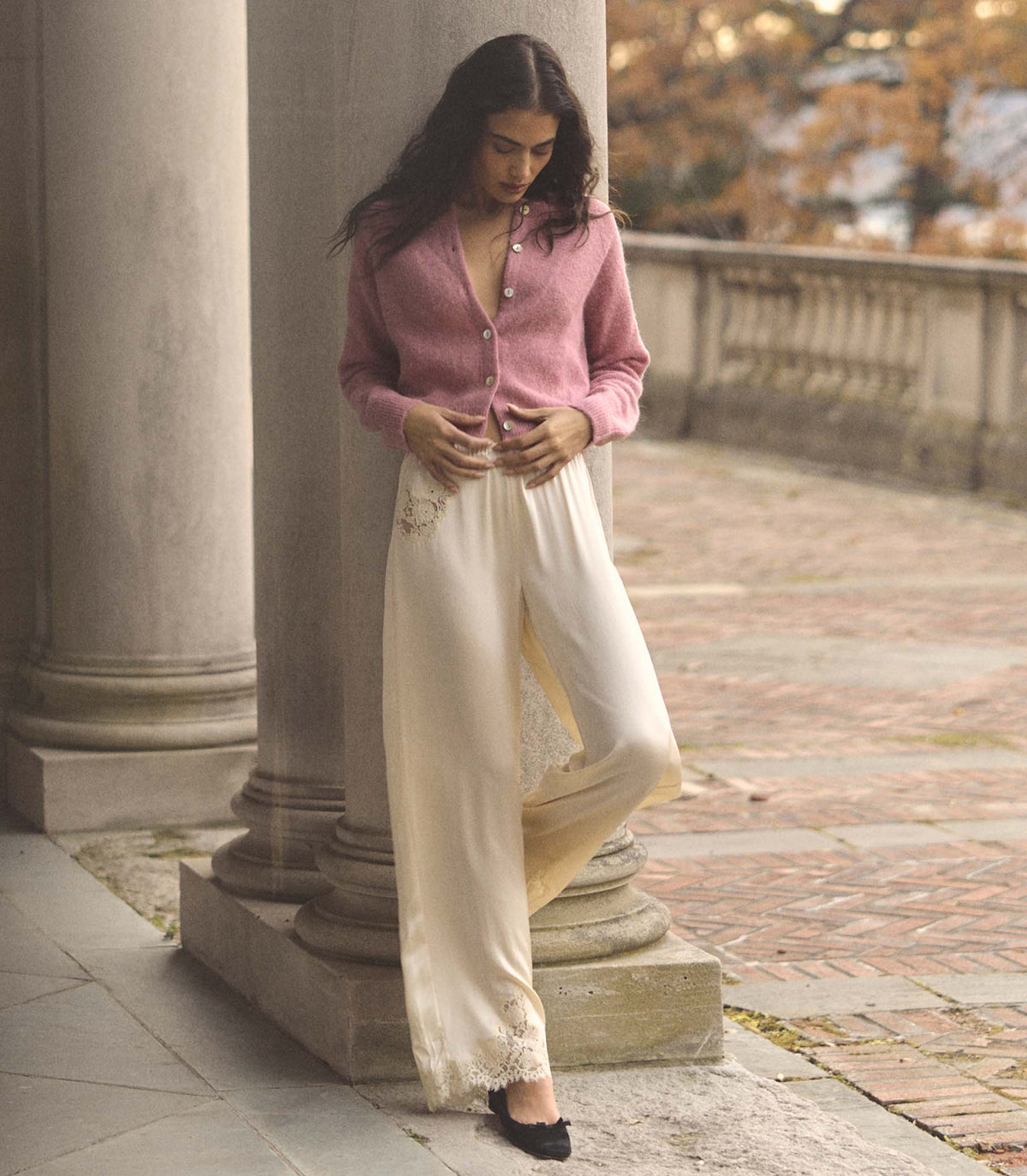 Woman in a pink cardigan and white pants standing on a stone path with columns in the background.