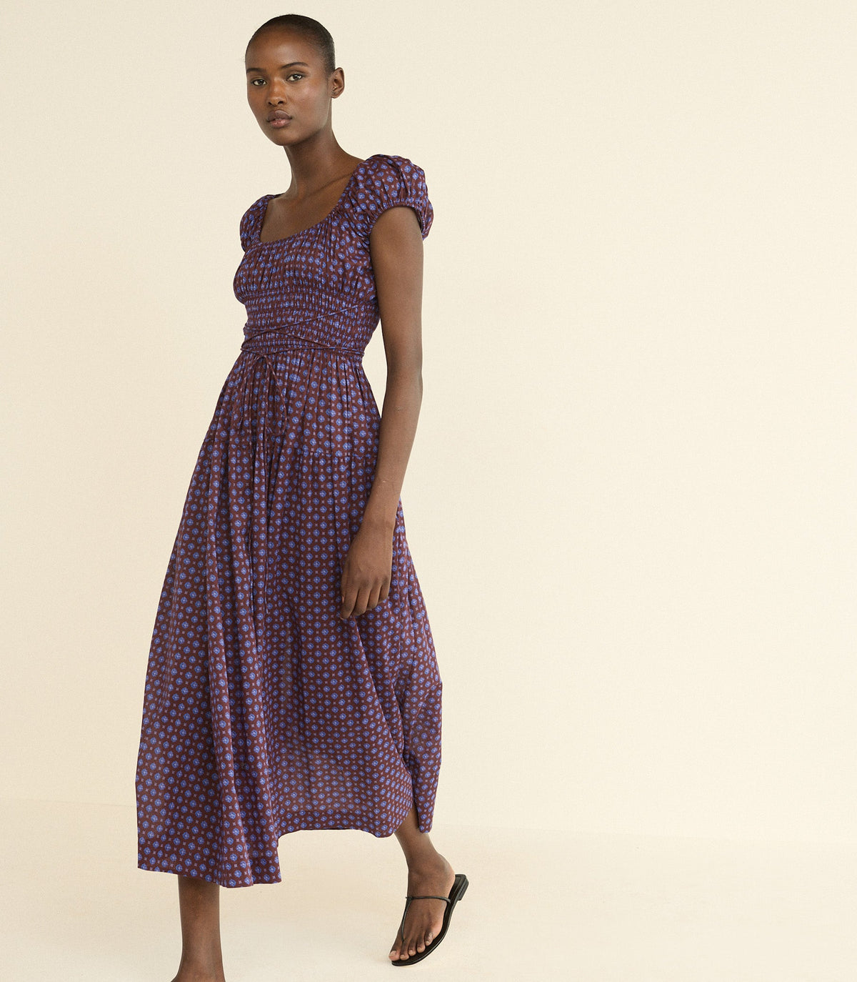 A woman modeling a midi-length purple dress with a small red and white foulard pattern, puffed cap sleeves, and a smocked bodice, set against a solid cream background.