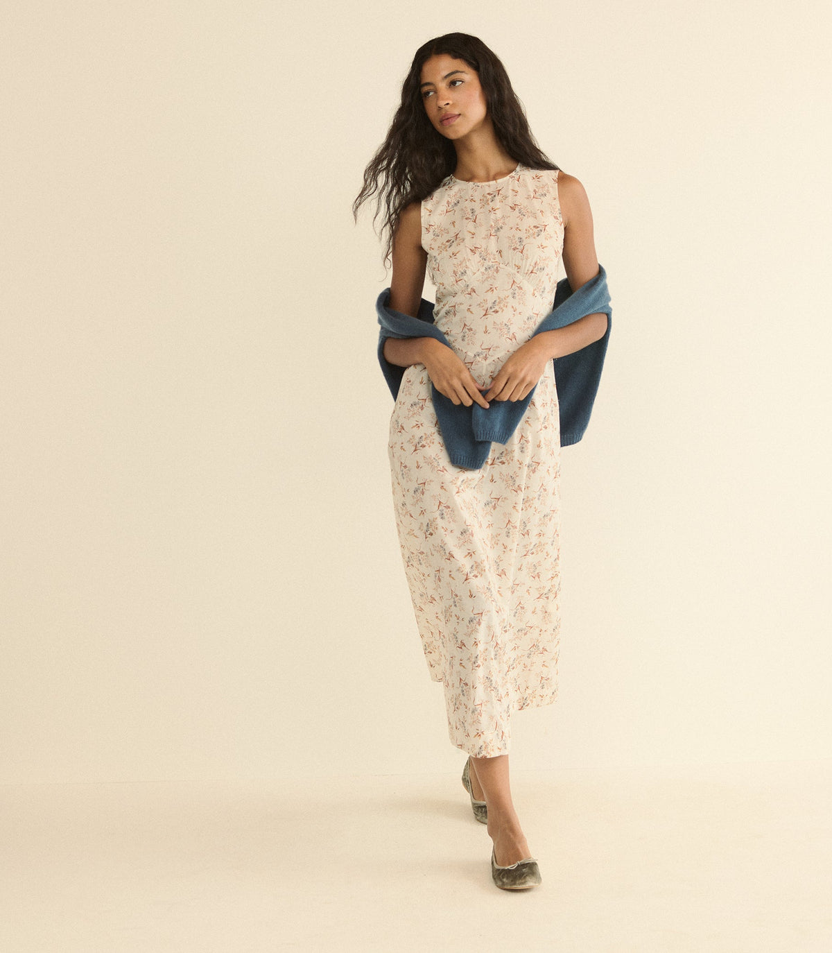 A woman modeling a sleeveless cream dress with a delicate floral print, styled with a draped blue shawl and metallic flat shoes, set against a solid warm beige studio background.