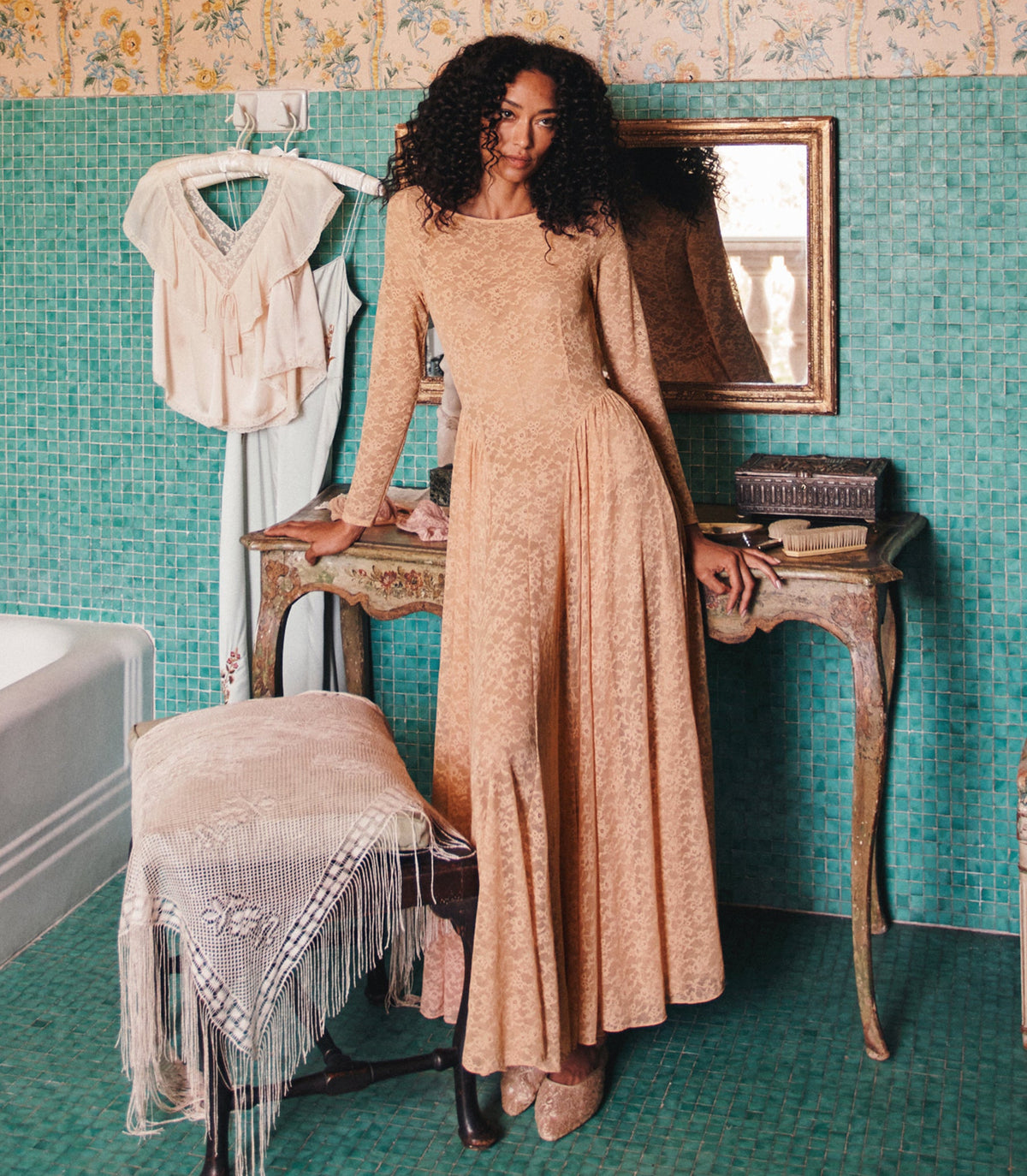 A woman with curly dark hair models a long-sleeved beige lace maxi dress in a vintage-style room with teal mosaic tile walls, leaning on a wooden table.