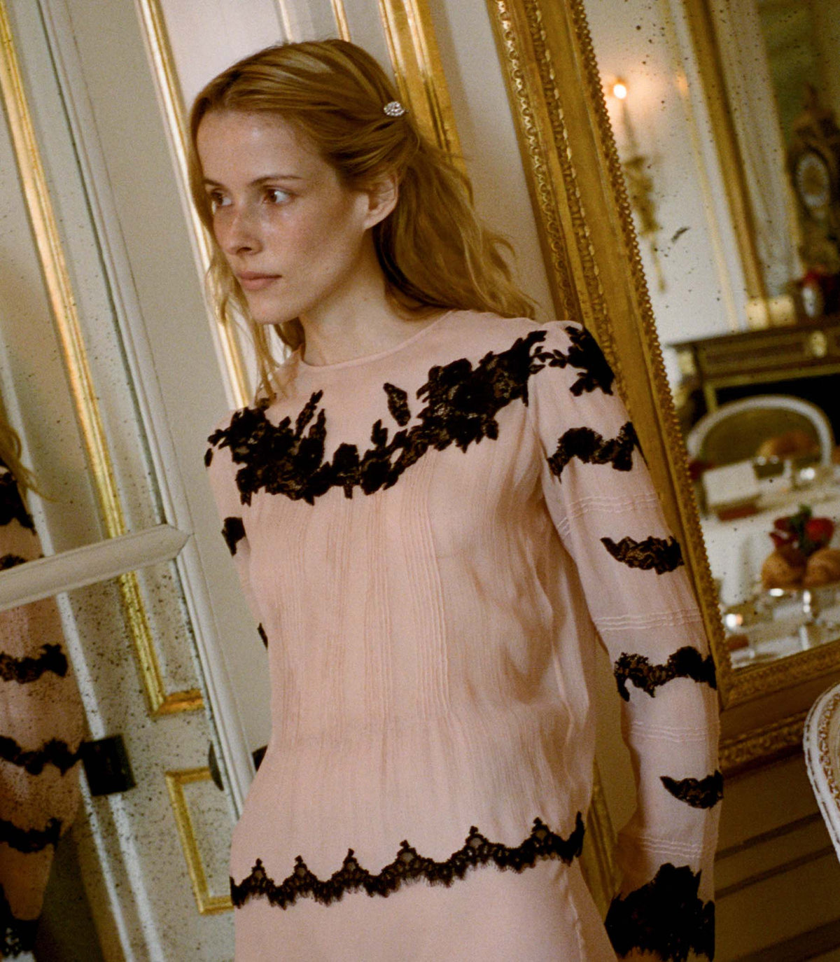Woman wearing a pink blouse with black lace details in an elegant room.