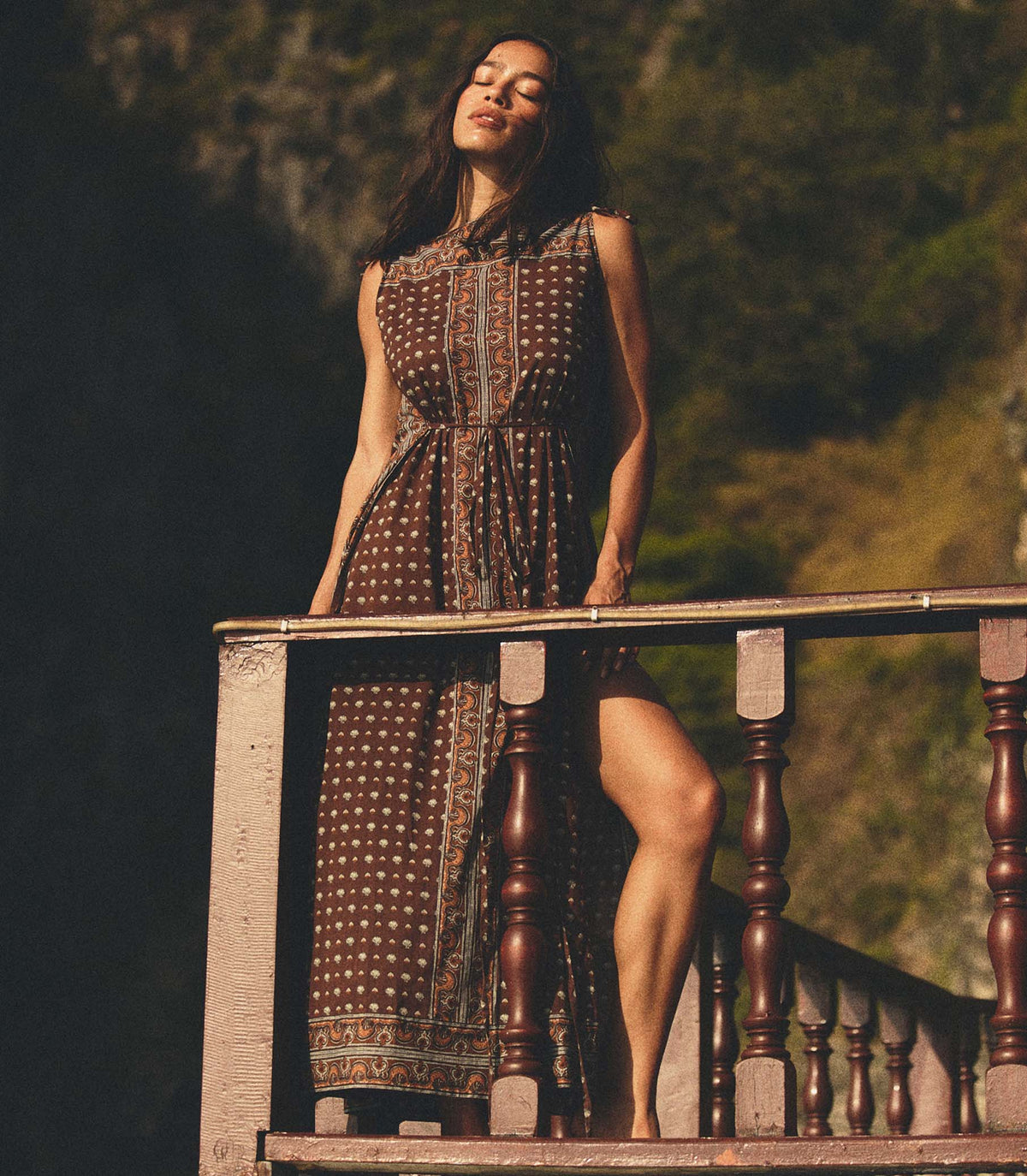 Woman in a patterned dress standing on a wooden railing with a blurred natural background