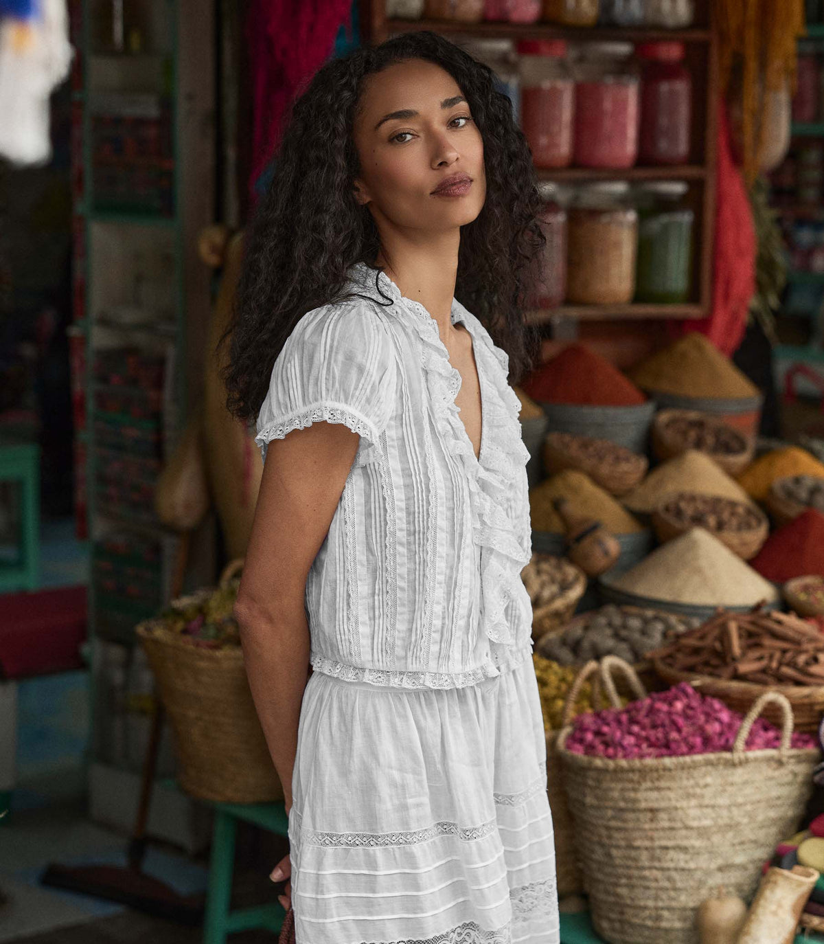 Woman in a white blouse standing in a market setting with various products and baskets.