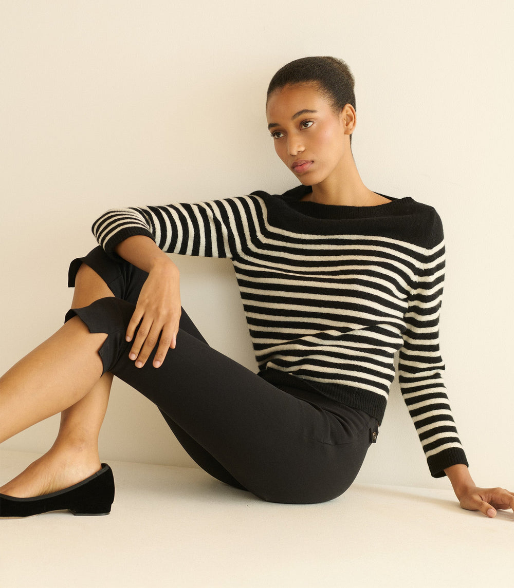 Woman wearing a black and white striped sweater and black pants sitting against a beige wall.