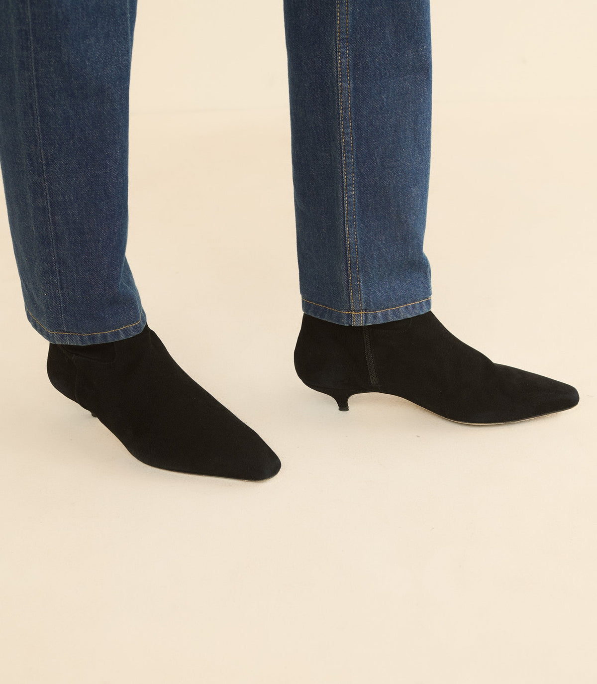 Black pointed-toe ankle boots with kitten heels, shown on a model wearing blue jeans against a plain beige background.