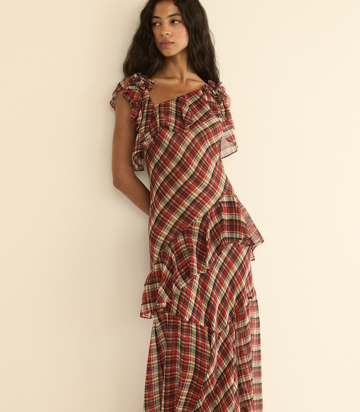 A woman wearing a red and cream plaid ruffled dress, posing against a plain beige wall.