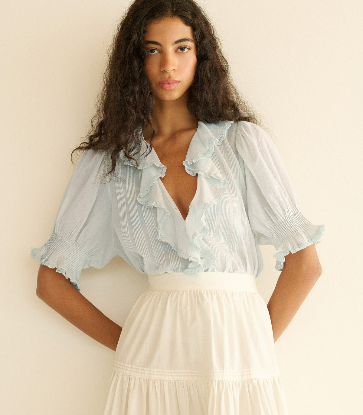 Woman wearing a light blue blouse with ruffles and a white skirt against a beige background