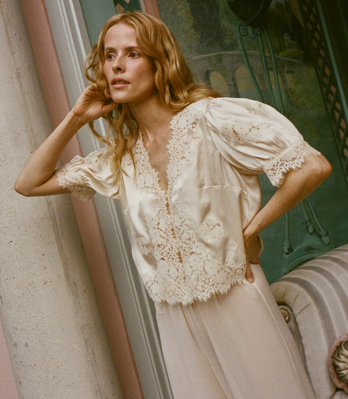 A woman wearing a cream-colored lace-trimmed silk blouse with puff sleeves and cream pants, posed in front of a vintage-style window.