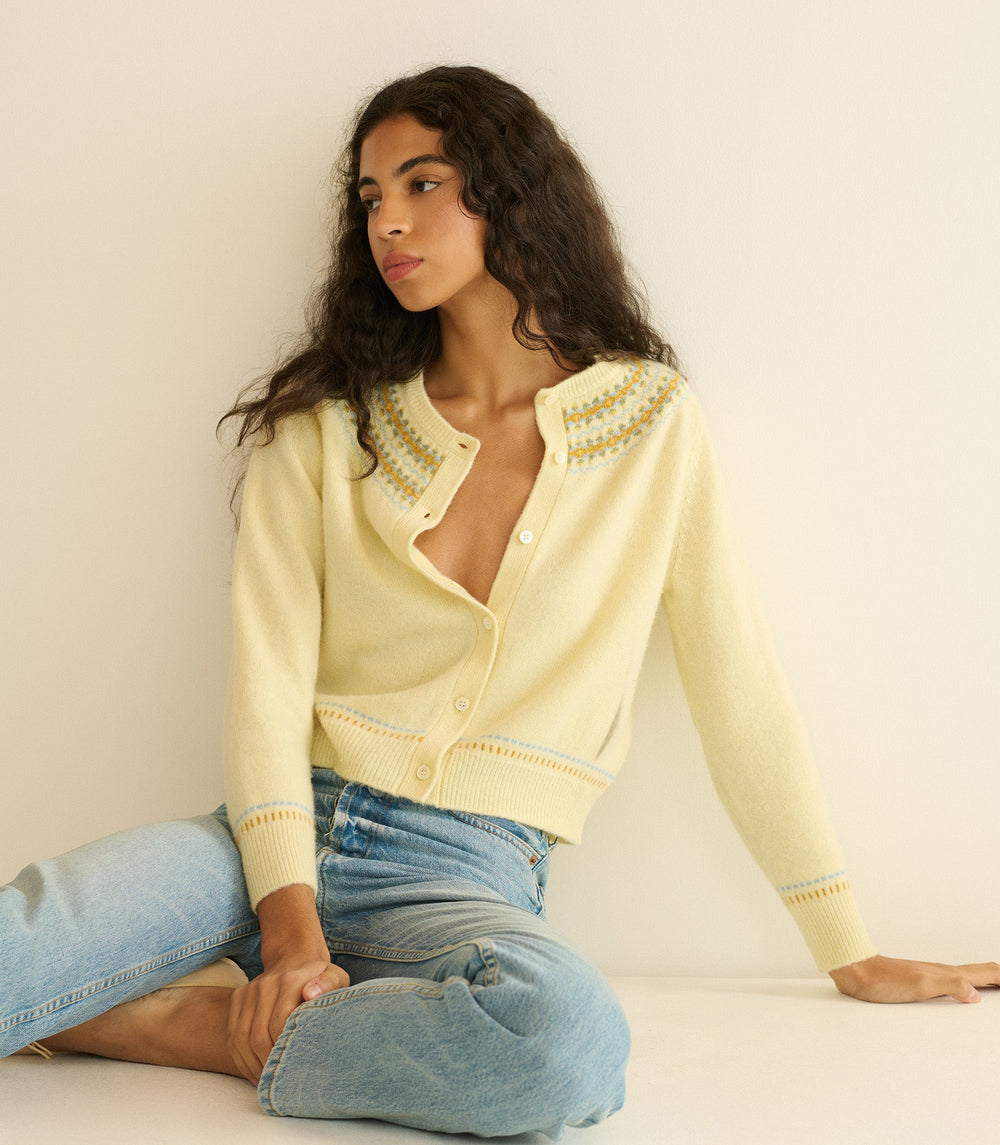 Woman wearing a light yellow cardigan with intricate patterns, sitting against a plain background.