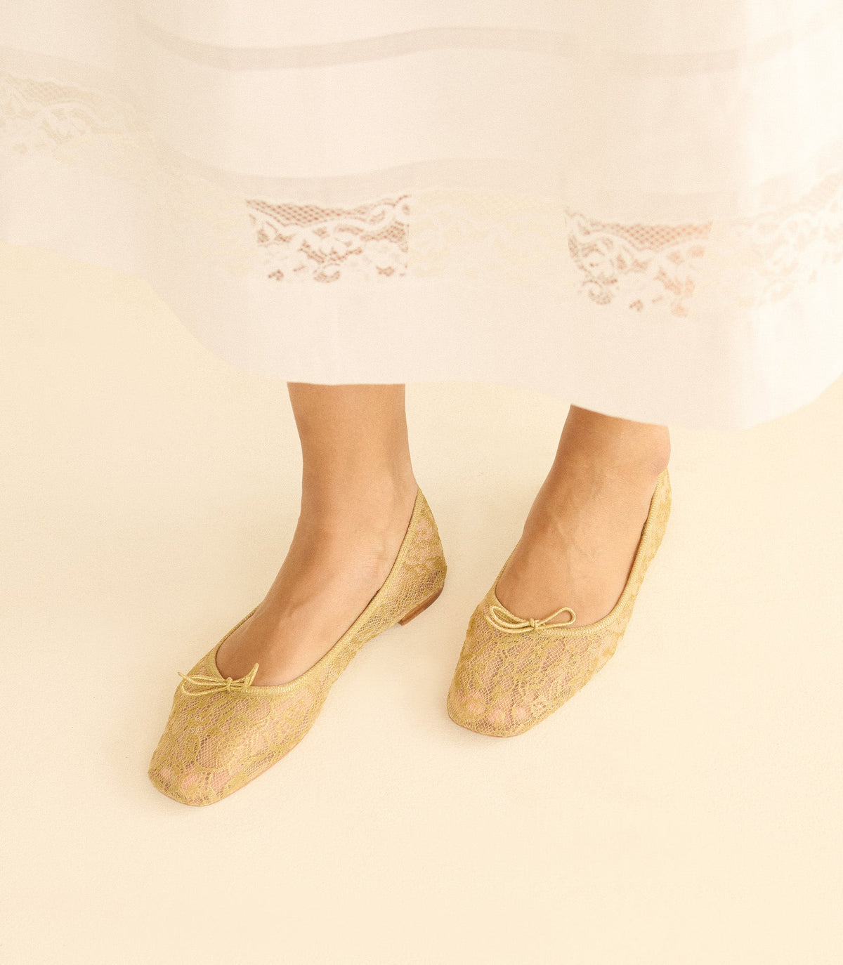 Gold lace ballet flats worn with a lace dress on a beige background